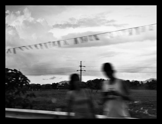 A black & white shot of a utility pole on a grass field, unintentionally obscured by motion blurred two people near the guardrails in the foreground. A woman seems to be waiting for a tricycle, while her son next to her is playing something in his phone.