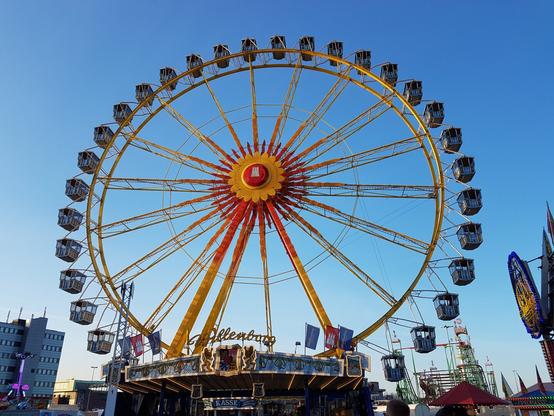 A large yellow Ferris wheel with red accents stands tall against a clear blue sky. The gondolas are visible, and the base of the ride is lit up with various lights. Other fairground attractions and buildings can be seen in the background.