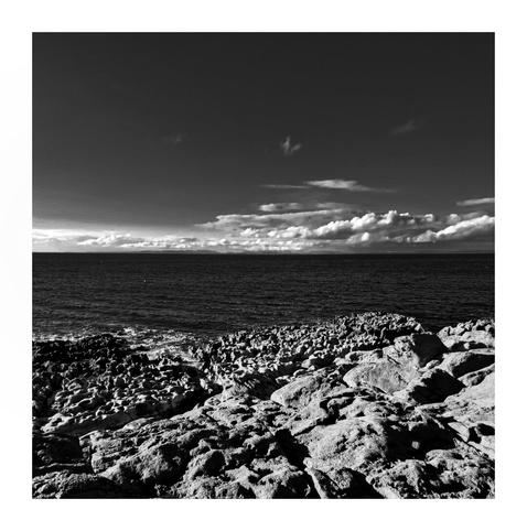 This is a black and white photo and it shows a rocky coastline in the foreground, which is a lifht grey colour and made of interesting irregular shapes. The ocean dark coloured ocean stretches out to the horizon. Above the water, there are scattered clouds beneath a mostly clear sky. The clouds are white and concentrate mostly on the left hand side of the photo close to the horizon.