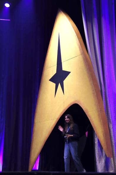 A person stands on stage, smiling and waving, next to a large yellow Starfleet insignia with a black star, illuminated by purple lights. Curtains drape in the background.
