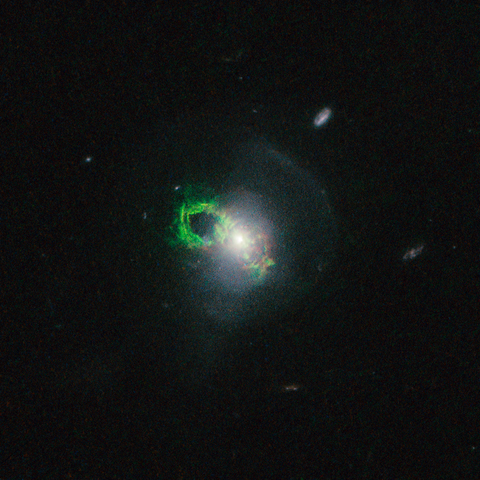 A Hubble Space Telescope image of the Teacup Galaxy, featuring a bright white core surrounded by swirling, faint blue gas. A vivid green, glowing ring of gas appears , shaped like a loop, is in the center left of the image. The surrounding space is dotted with distant galaxies and stars.