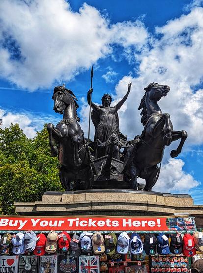 A low-angle shot showcases the bronze statue of Boudica, the warrior queen of the Iceni, standing regally in her chariot. Two powerful horses pull the chariot, their muscles defined and appearing to be in motion. Boudica faces forward, a spear held upright in her right hand, her left arm raised in a commanding gesture. She is adorned in a flowing gown and a crown or headdress, signifying her royal status. The statue is set against a backdrop of a bright blue sky dotted with fluffy white clouds, suggesting a sunny day in London. In the immediate foreground, slightly blurred, is a souvenir stand. A bright red banner at the top of the stand proclaims "Buy Your Tickets Here!". Below the banner, an assortment of London-themed souvenirs are displayed, including baseball caps emblazoned with "LONDON", keychains, and other small trinkets, indicating the statue's location as a popular tourist attraction near Westminster Bridge.