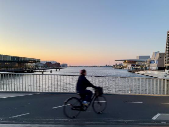 Fahrrad fährt vor Sonnenuntergang über eine Brücke, im Hintergrund Meer und Stadt