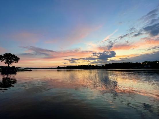 Sunset photo of peach and gray clouds over a reflective lake, with a tree on the shore on the left-hand side.