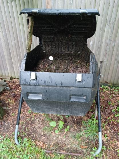 A large, black plastic compost tumbler with the lid propped open by a log.  Inside is a mix of grass clippings and shredded leaves, partially decomposed and greenish-brown.  
There is a dial-type compost thermometer stuck in to the contents.