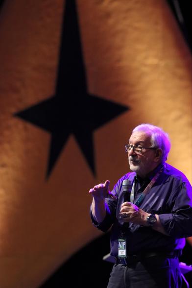 A speaker holds a microphone and gestures while presenting on stage in front of a large star backdrop. The setting appears to be a conference or event.