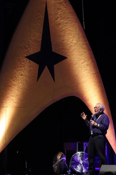 A speaker stands on stage, gesturing while holding a microphone. Behind them is a large backdrop featuring a yellow emblem with a black star, reminiscent of the Starfleet logo from "Star Trek." The setting is a dimly lit auditorium with fans visible.