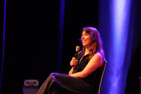 A woman with long hair sits on a chair holding a microphone, smiling brightly. She is wearing a black sleeveless top and dark jeans, with soft blue and purple stage lighting in the background.