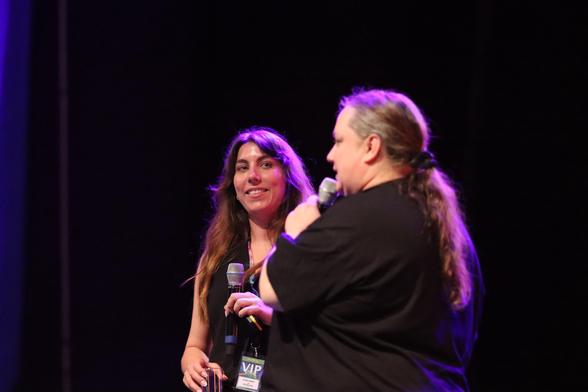 A woman and a person with long hair are speaking on stage, both holding microphones. The woman is smiling and appears engaged in the conversation. The background features stage lighting, adding a vibrant atmosphere to the setting.