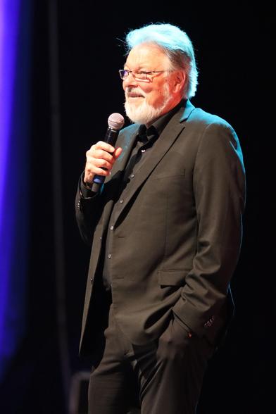 A man with gray hair and a beard is standing on stage, holding a microphone and smiling. He is dressed in a black suit and shirt, with the background featuring soft lighting.