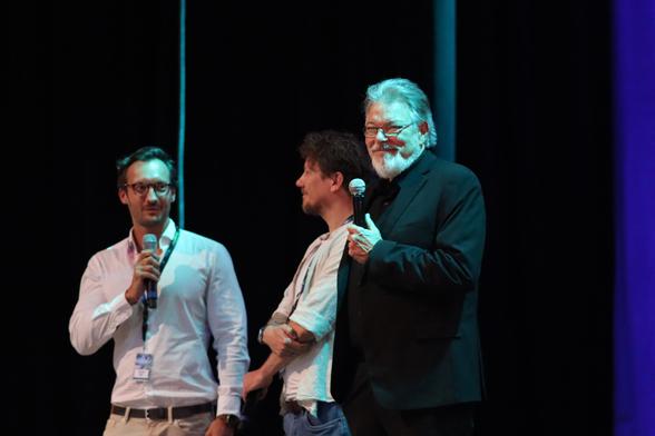 Three individuals are on stage, with one holding a microphone and speaking. The person on the right has gray hair and a beard, dressed in a black suit. The middle individual has curly hair and is dressed casually. The background is dark.