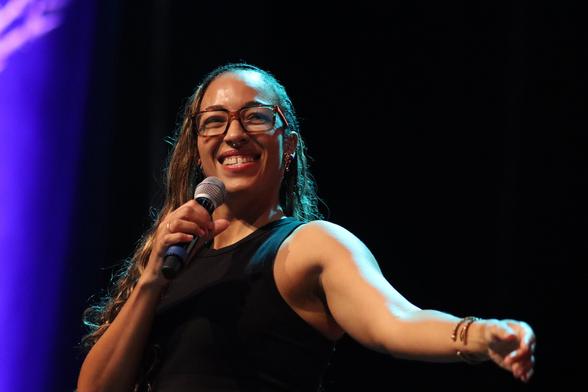 A person with long hair, wearing glasses, smiles while holding a microphone. They are speaking or performing on stage, with a dark background and colorful lighting.
