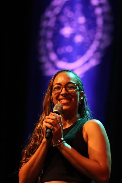 A smiling person with long hair and glasses holds a microphone, standing under vibrant stage lighting. The background features a decorative emblem or logo.