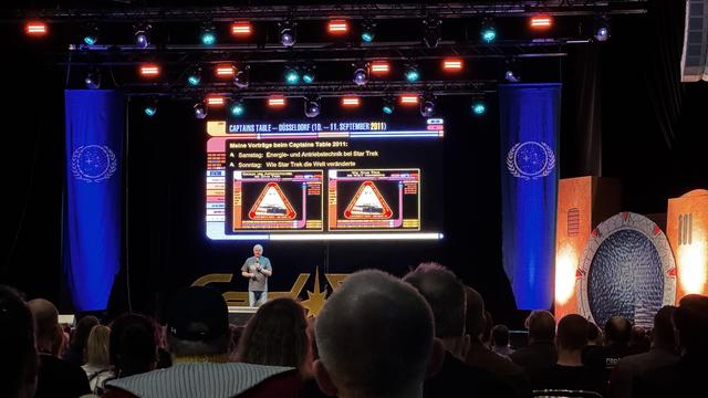 An event speaker presents at a conference, discussing Star Trek topics. A large screen displays information in German about the presentations, including "Energy and Propulsion Technology in Star Trek" and "How Star Trek Changed the World."