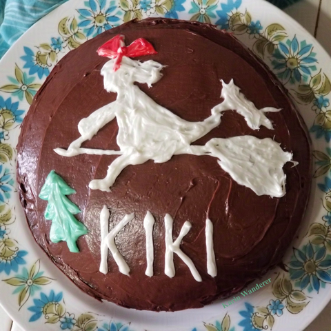 Photo of a chocolate cake decorated with chocolate frosting. Drawn in white frosting is a witch riding a broom, with a cat hanging on to her. The witch is wearing a red hair bow and flying over a green tree, as well as the name "Kiki" in white.