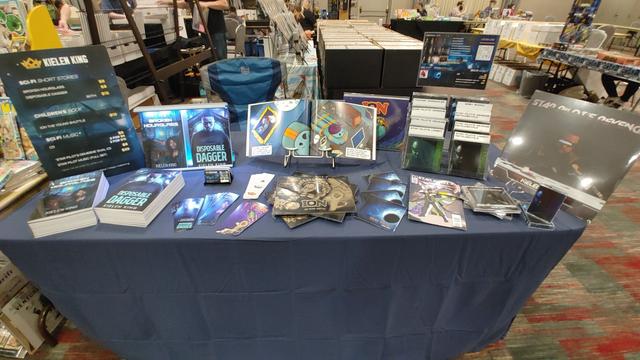 Convention table set up with books, bookmarks, business cards, CDs, cassettes, and vinyl records for sale