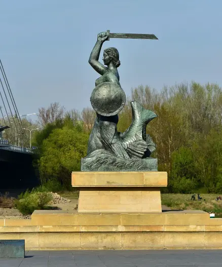 The image depicts a large statue of a woman holding a sword and a shield. She has a fish tail, suggesting a mermaid figure. The statue is situated on a pedestal, and there are trees and a bridge in the background. The setting appears to be outdoors, likely near a body of water.

Image Credits: Wikimedia / Adrian Grycuk / CC BY-SA 3.0 pl