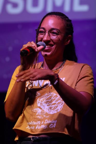 A person is talking into a microphone on stage, wearing glasses and a yellow graphic t-shirt. They have braided hair and are smiling. The background features a purple hue with text partially visible.