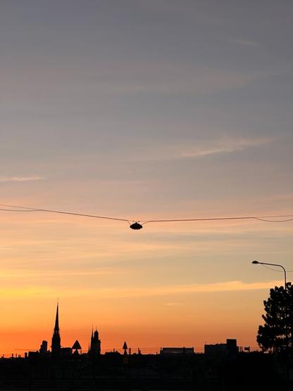 a skyline, with a church-like spire, some other buildings, a tree and a few signs at various levels of depth, silhouetted by a radiant sunset. An overhead street light of suspended in the middle of the frame on a cable running horizontally.