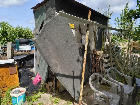 A shed with a panel at an angle of about 30 degrees. There is a support and a bit of guttering. Two plastic chairs, a wood pallet, and a bucket are visible.