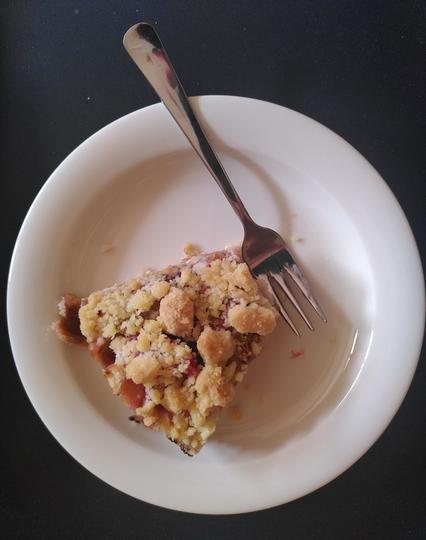 A piece of rhubarb crumble cake on a white plate on a black background. A fork on the plate, one bite is already missing.