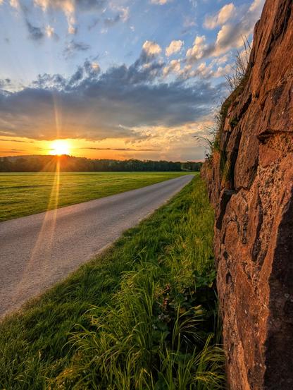 A ground-level, wide-angle shot captures a country road stretching into the distance at sunset. The low sun, positioned on the left horizon, casts long rays across a lush green field to the left of the road. The sky is a mix of blues and whites, with streaks of orange and yellow near the setting sun. To the right of the road, a weathered, reddish-brown stone wall runs along the frame's edge, with tall green grass growing at its base. The road itself is a light grey, and it curves slightly to the right as it recedes towards the line of trees on the horizon.