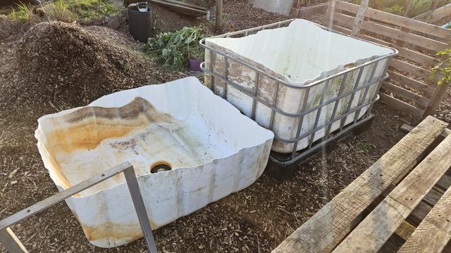 A cut in half IBC, bottom half (a larger half TBH) white plastic inner in the cut down cage, the top half of rhe plastic is sat to the left of this without any frame. Both are open side upwards.