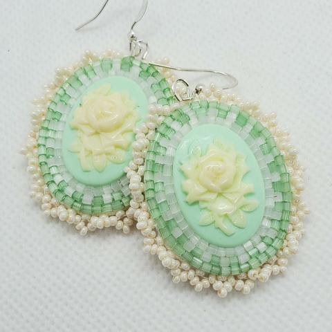 an elegant pair of beaded earrings. the centerpiece is a cream colored rose on a pastel green background surrounded by clear, light green, and pearlescent beads to accentuate.