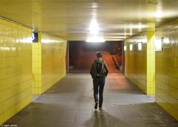 Eine Unterführung führt zu den Gleisen eines kleinen Bahnhofs. Wände und Decke sind gelb gefliest. Mitten im Tunnel geht eine Reisende mit einem kleinen Rucksack auf dem Rücken davon. /
An underpass leading to the tracks of a small train station. The walls and ceiling are tiled yellow. In the middle of the tunnel, a traveler walks away with a small backpack on her back.