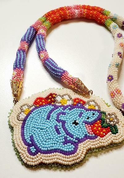 a beaded medallion made by kiamichi of a baby hippo in blue eating a strawberry in a field of strawberries and their flowers. the necklace that holds the medallion is multiple colors with floral accents