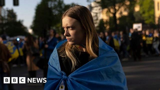 A woman stands looking to her side at a protest wrapped in a blue Ukraine flag.