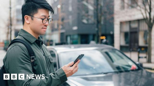 Man holding a phone stands next to a black car in a street.