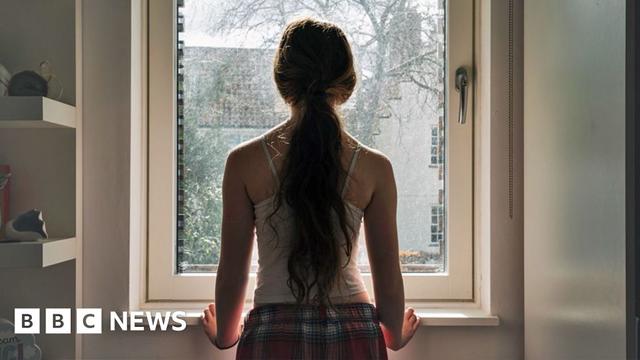 Stock image of a girl with long hair tied in a ponytail stands next to a window and looks outside, wearing a vest and checked pyjama bottoms.