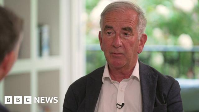 Robert Harris sits in a bright room with a balcony and trees visible in the background. He wears a dark blazer and a white shirt.