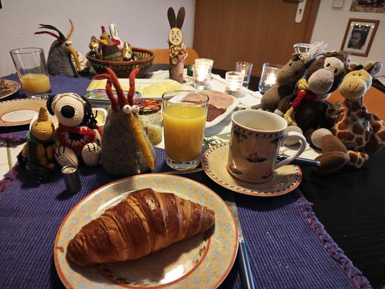 Frühstückstisch mit lila Tischset, Sally wärmt das Frühstücksei, die letzte Osterdeko steht noch, vorne auf dem Teller ein Croissant, Orangensaft im Glas, auf dem Tisch sitzen zwischen den Wurst und Käseplatten diverse Plüsch und Häkeltiere. 