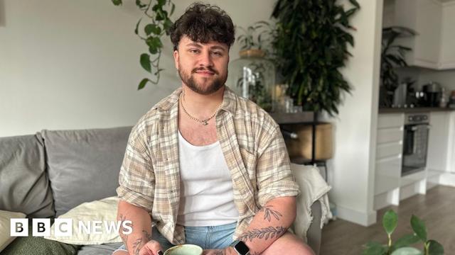 A man with dark brown hair and a beard sits on a sofa smiling at the camera. He's wearing a checked beige shirt over a white vest and has tattoos