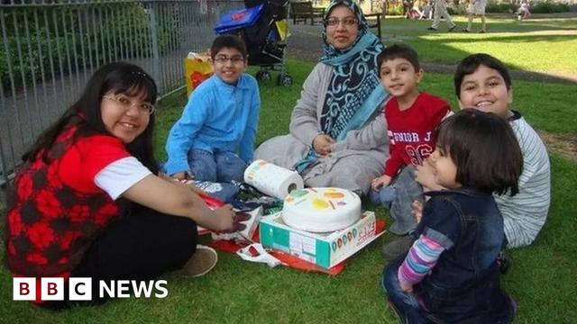 Sabah Usmani and her five children smile at the camera as they sit around a birthday cake in a park. Dr Usmani is wearing a blue headscarf and grey cardigan.