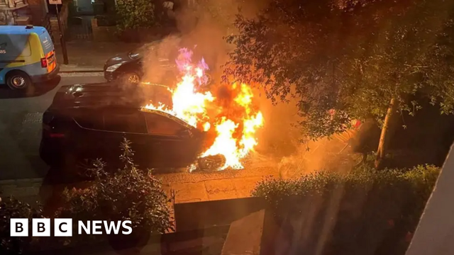 The front half of a car parked on a residential street is seen engulfed in flames.