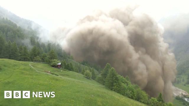 A large cloud generated by rock and mud rises over the green slope of a valley