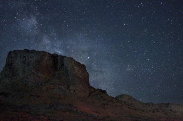 A wide-angle, low-light photograph captures a rugged mountain formation against a vast, star-filled night sky, with the Milky Way faintly visible. The mountain occupies the left half of the frame, its dark, textured face rising sharply. The right half of the image is dominated by the night sky, a deep blue-black adorned with countless pinpricks of light from stars. A wide-angle, low-light photograph captures a rugged mountain formation against a vast, star-filled night sky, with the Milky Way faintly visible. The mountain occupies the left half of the frame, its dark, textured face rising sharply. The right half of the image is dominated by the night sky, a deep blue-black adorned with countless pinpricks of light from stars.