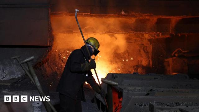 A steel worker at British Steel site in Scunthorpe in April 2025 - the picture is largely dark, which dramatically frames one worker standing in front of a furnace, his outline lit up by the furnace behind him - he is wearing a protective helmet and goggles, and holding what looks to be a long pipe 