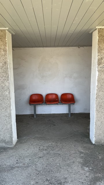 Drei rote Stühle, die an einer weißen Wand unter einem Unterstand mit grauem Betonboden stehen.