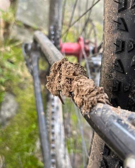 Nahaufnahme des Hinterrads eines Fahrrads mit Schlamm auf dem Reifen, fokussiert auf einen Schlammklumpen auf dem Rahmen in der Nähe der Zahnräder mit einem unscharfen grünen Hintergrund.