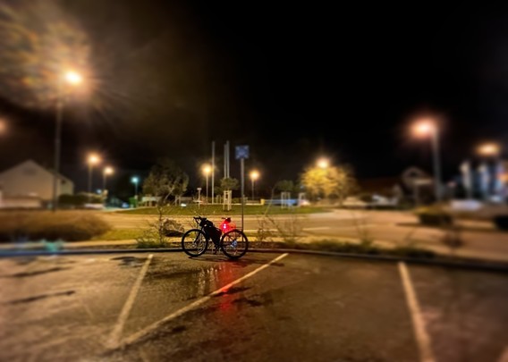 Ein Fahrrad mit rotem Rücklicht, das auf einem nächtlichen Parkplatz geparkt ist, mit Straßenlaternen und einem unscharfen Hintergrund.