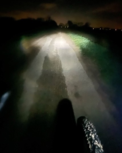 Ein unbefestigter Weg, der nachts von einer Fahrradlampe beleuchtet wird und auf dem der Schatten eines Fahrradreifens zu sehen ist.