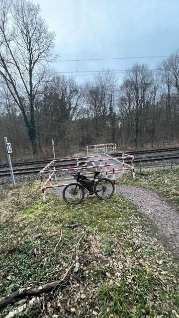 Ein Fahrrad, das auf einem unbefestigten Weg in der Nähe von Bahngleisen gegen eine Absperrung gelehnt ist, mit Bäumen im Hintergrund und bedecktem Himmel.