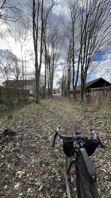 Ein Fahrrad auf einem mit Laub bedeckten Weg, der von kahlen Bäumen gesäumt ist, mit Schuppen zur Rechten und einem klaren Himmel darüber.
