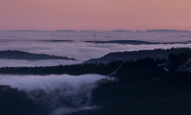 View from a Black Forest summit at dusk during inversion weather conditions. The highest peaks emerge from a sea of fog. In the center of the image, a single wind turbine protrudes from the mist. Far in the distance, the Alpine range is visible. Some mountain peaks are distorted and partially elevated in a pillar-like manner, due to the effect of mirages.