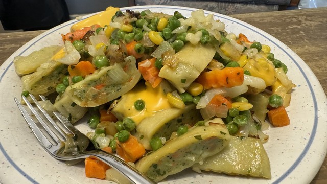 Ein Teller Maultaschen mit geschmolzenem Käse, Erbsen, Karotten, Mais und gewürfelten Zwiebeln, mit einer Gabel an der Seite.