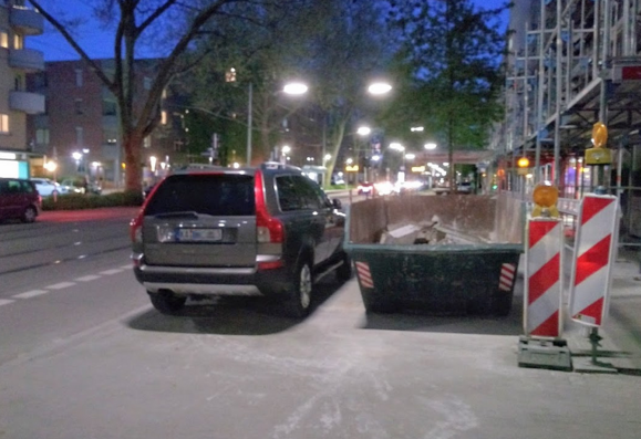 Nachtaufnahme: parkender PKW blockiert exakt die Gehwegrestbreite, die neben Gerüst und Bauschuttmulde noch verbleibt.